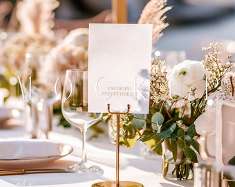 May include: A white card with the text "PSD & JPG SMART OBJECT" on a gold metal stand. The stand is on a white tablecloth with a glass of water and a plate. There are white flowers in a vase behind the stand.