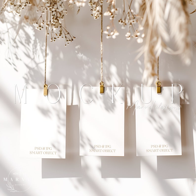 May include: Three white cards hanging from twine with gold clips against a white wall. The cards are blank and have the text "PSD & JPG SMART OBJECT" printed on them. The cards are surrounded by dried flowers and greenery.