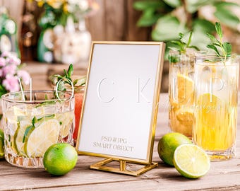 Può includere: Un cartello con cornice dorata con le lettere "CK" e il testo "PSD & JPG SMART OBJECT" è posto su una superficie di legno. Accanto ci sono bicchieri di cocktail con fette di lime e arancia e lime freschi. Composizioni floreali sullo sfondo.
