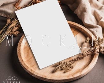 May include: A blank white card mockup on a wooden plate with dried flowers and a beige fabric in the background. The text "MOCKUP" is written in white on the plate.