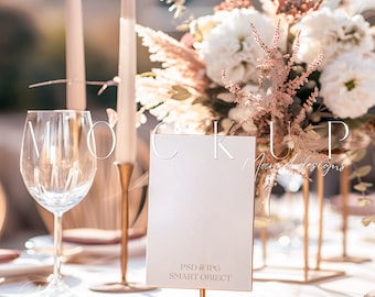 May include: A white card with gold trim sits on a gold stand on a white tablecloth. The card has the text "PSD & JPG SMART OBJECT" printed on it. The card is surrounded by a white and pink floral arrangement, a wine glass, and gold candlesticks.
