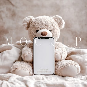 Può includere: Un orsacchiotto beige che tiene in mano uno smartphone bianco con uno schermo nero. Il telefono ha il testo "PSD & JPG SMART OBJECT" sullo schermo. L'orsacchiotto è seduto su una coperta bianca.