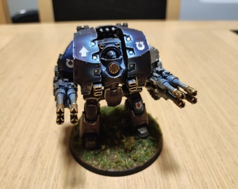 May include: A dark blue and black painted miniature robot with gold accents and a white arrow symbol on the front. The robot is standing on a green base.