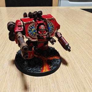 May include: A red and gold miniature war machine with multiple weapons, standing on a black base with a lava-like design. The model features intricate details and is likely a collectible item or a piece for tabletop gaming.