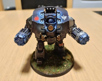 May include: A black and blue robot with a red maple leaf on its chest and a white symbol on its shoulder. The robot has two large guns on its arms and is standing on a green base.