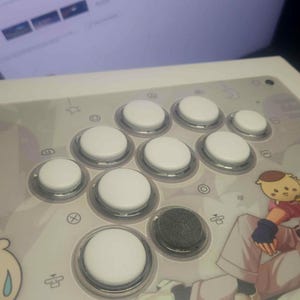May include: A white arcade controller featuring a layout of circular white buttons and one black button. The surface is decorated with a graphic illustration of a character with a small yellow creature on their shoulder and a crying cartoon face.