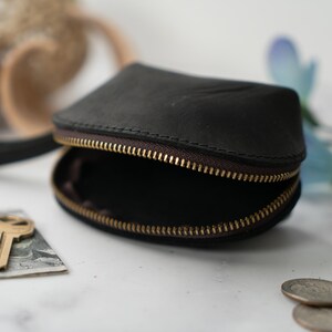 May include: A small, black leather coin purse with a zip closure and a short handle. The purse is open, revealing the empty interior. A key and a dollar bill are lying on a white surface next to the purse.