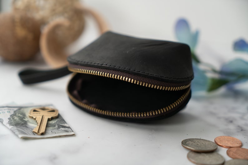 May include: A small, black leather coin purse with a zip closure and a short handle. The purse is open, revealing the empty interior. A key and a dollar bill are lying on a white surface next to the purse.