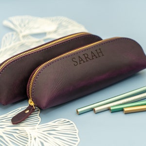 May include: Two purple leather pencil cases with gold zippers. The larger case has the name "SARAH" embossed on the front. The cases are on a white background with a few pencils in the foreground.