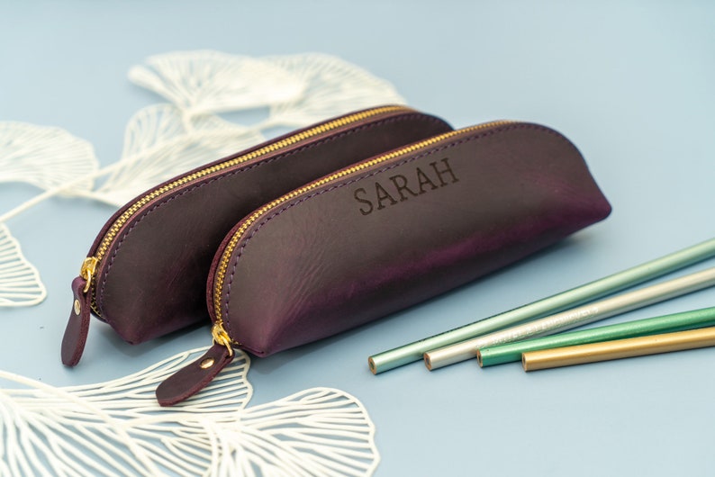 May include: Two purple leather pencil cases with gold zippers. The larger case has the name "SARAH" embossed on the front. The cases are on a white background with a few pencils in the foreground.