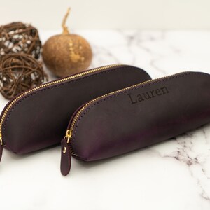 May include: Two purple leather pencil cases with gold zippers. The larger case is plain, while the smaller case has the name "Lauren" embossed on it.