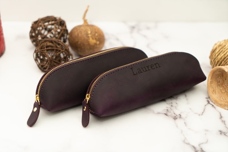 May include: Two purple leather pencil cases with gold zippers. The larger case is plain, while the smaller case has the name "Lauren" embossed on it.