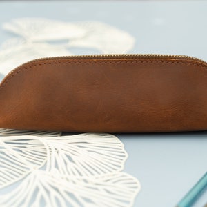 May include: A brown leather pencil case with a zipper closure. The case is on a light blue surface with white paper decorations.