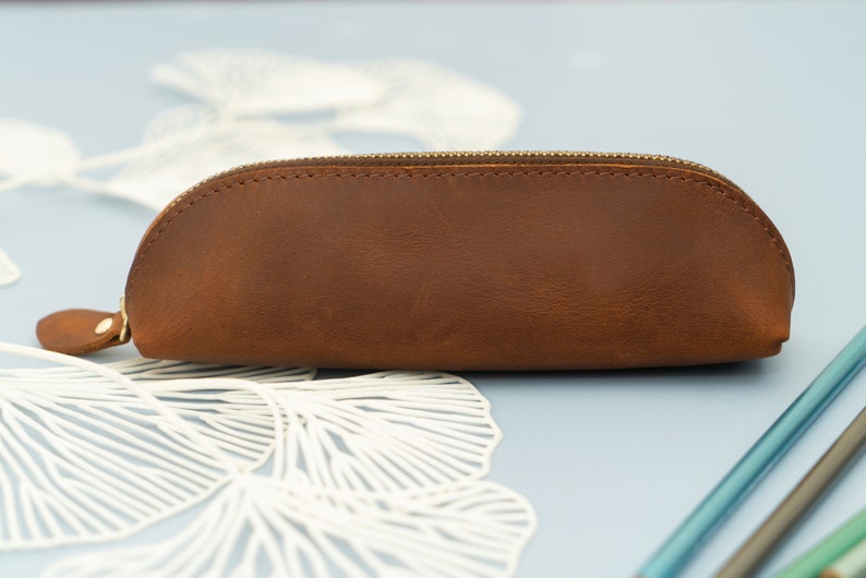 May include: A brown leather pencil case with a zipper closure. The case is on a light blue surface with white paper decorations.
