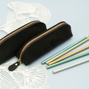 May include: Two black leather pencil cases with gold zippers. The larger case has the word "Charlie" embossed on it. Five metallic pencils are lying on a white background.