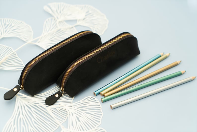 May include: Two black leather pencil cases with gold zippers. The larger case has the word "Charlie" embossed on it. Five metallic pencils are lying on a white background.