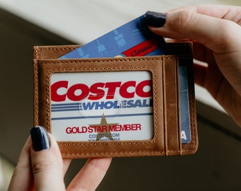 May include: A brown leather cardholder with a clear window displaying a Costco Wholesale membership card. A blue credit card is being inserted into a slot. The cardholder has visible stitching.