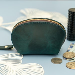 May include: A teal green leather coin purse with a zipper closure and a short strap. The purse is sitting on a light blue surface with white decorative items and coins.