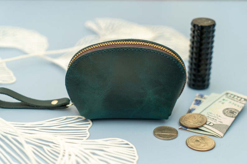 May include: A teal green leather coin purse with a zipper closure and a short strap. The purse is sitting on a light blue surface with white decorative items and coins.