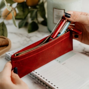 May include: A red leather pencil case with a gold zipper is open, revealing two pens inside. The case is held in a hand.