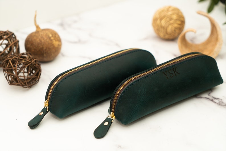 May include: Two green leather pencil cases with gold zippers. The smaller case has the initials "YSK" embossed on the front.