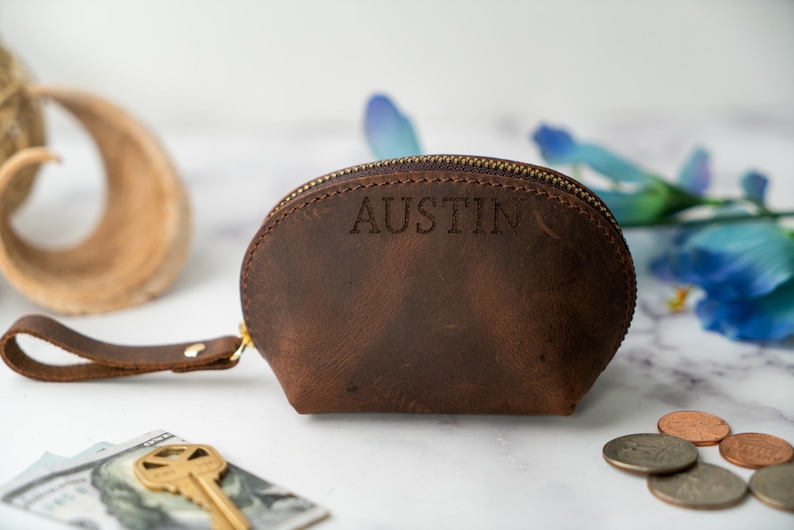 May include: A brown leather coin purse with a zip fastening and a small strap. The purse is personalised with the name "Austin".