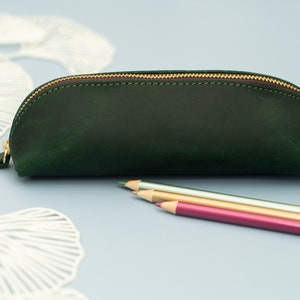 May include: A dark green leather pencil case with a gold zipper. The case is closed and sitting on a light blue surface.
