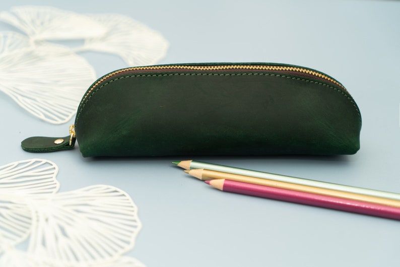 May include: A dark green leather pencil case with a gold zipper. The case is closed and sitting on a light blue surface.