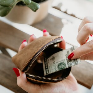 May include: A brown leather coin purse with a zip fastening, open to reveal a dollar bill inside. The purse is held in a hand with red nail polish.