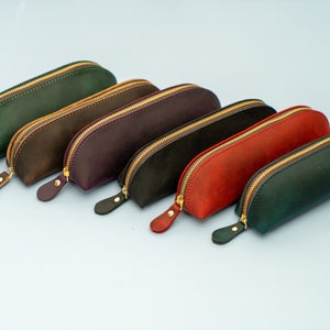 May include: Six leather pencil cases in different colors, including green, brown, purple, black, red, and dark green. Each case has a gold zipper and a curved shape.
