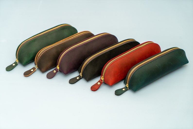 May include: Six leather pencil cases in different colors, including green, brown, purple, black, red, and dark green. Each case has a gold zipper and a curved shape.
