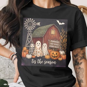 May include: A black t-shirt featuring a graphic print of a red barn, a windmill, pumpkins, and three ghosts. A wooden sign reads PUMPKINS HAY RIDES CORN MAZE. The text tis the season is printed at the bottom of the graphic in a white cursive font.