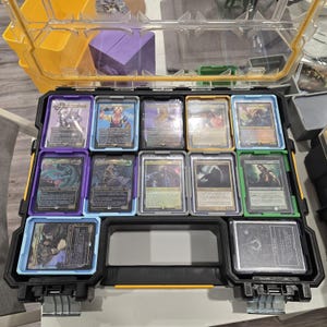 May include: A black and yellow plastic storage case with a clear lid, filled with trading cards in protective sleeves. The cards feature colourful illustrations and text, organised in rows within the case.