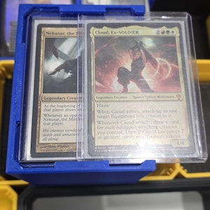 Könnte beinhalten: Ein blauer Kartenhalter aus Kunststoff mit zwei Magic: The Gathering Sammelkarten. Die Karten sind in Schutzhüllen. Eine Karte zeigt eine dunkle Kreatur, die andere einen menschlichen Soldaten. Der Halter befindet sich in einem größeren Aufbewahrungsbehälter.