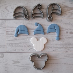 May include: An assortment of cookie cutters and cookies in the shape of Santa hats and a Mickey Mouse head. The Santa hat cutters are gray, and the hats are light blue. The Mickey Mouse head is white, and the cutter is gray.