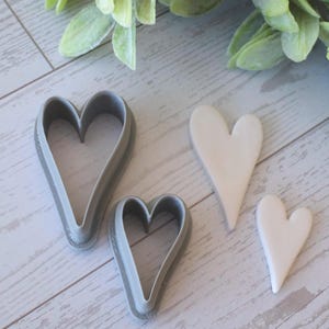 Puede incluir: Una colección de cortadores de galletas en forma de corazón y corazones de cerámica. Dos cortadores de plástico gris de diferentes tamaños están junto a dos corazones de cerámica blancos. El fondo incluye follaje verde sobre una superficie de vetas de madera clara.