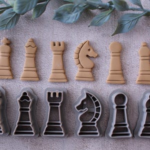 Puede incluir: Un conjunto de cortadores de galletas con forma de piezas de ajedrez: rey, reina, torre, caballo, alfil y peón. Los cortadores son de color beige y gris, con una hoja verde decorativa en el fondo.