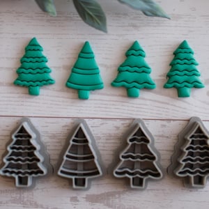 以下が含まれることがあります： 明るい木製の表面に、4つの緑色のクリスマスツリー型のクッキーカッターと4つの灰色のツリー型のクッキーカッターが配置されています。緑の木は無地で、灰色の木は内部に切り抜かれたデザインがあります。