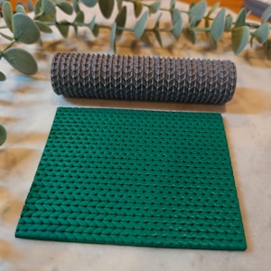 Könnte beinhalten: Eine dunkelgraue, strukturierte Rolle und ein grünes, strukturiertes Quadrat. Die Rolle hat ein sich wiederholendes Muster aus abgewinkelten Linien. Das grüne Quadrat hat ein sich wiederholendes Muster aus kleinen, erhabenen Punkten. Die Artikel befinden sich auf einer weißen Oberfläche mit grünem Laub im Hintergrund.