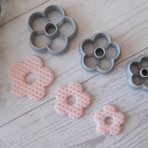 May include: An assortment of flower-shaped cookie cutters and pink floral-patterned clay pieces. The cutters are gray plastic, and the clay pieces have a textured surface. The items are arranged on a light wood-grain surface.