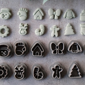 May include: A collection of Christmas-themed cookie cutters in various shapes, including a snowman, mitten, house, bow, sweater, Santa, wreath, reindeer, candy cane, gingerbread man, tree, and stocking. The cutters are white with black outlines.