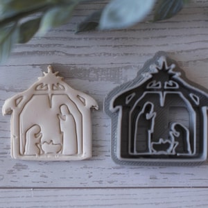 Puede incluir: Dos cortadores de galletas con forma de escena de la natividad. Uno es una galleta beige y el otro es un cortador de plástico gris. La escena de la natividad incluye a María, José y el niño Jesús en un pesebre, dentro de un establo.