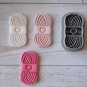 May include: A collection of yarn-themed cookie cutters in various colors. The cutters are shaped like yarn balls with a heart detail. One is a gray cutter, and the others are white and shades of pink. Ideal for baking themed cookies.