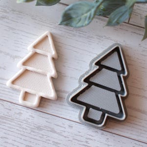 May include: Two Christmas tree-shaped cookie cutters. One is a light beige colour, and the other is grey with a black outline. The cutters are made of plastic and sit on a light wood-grain surface.