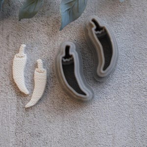 May include: Two white chili pepper-shaped soap bars and two gray chili pepper-shaped molds. The soap bars have a textured surface. The molds have a dark gray interior and a light gray outline. The items are on a light gray textured surface.