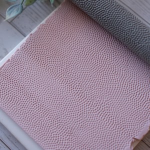 May include: A close-up of pink and grey textured leather materials. The pink leather is flat with a dotted pattern. A roll of grey leather with a similar pattern is on top. The materials are on a white surface.