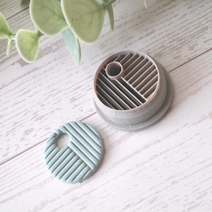 May include: A gray, circular clay cutter and a matching clay piece with a woven pattern. The cutter has a cylindrical shape with a grid design. The clay piece is light blue-green with a hole. The items are on a white wooden surface.