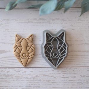 May include: A geometric wolf head cookie cutter and a baked cookie. The cookie cutter is gray plastic, and the cookie is light brown. The wolf head design is made of angular lines, creating a modern, artistic look. The cookie is on a light wood surface.