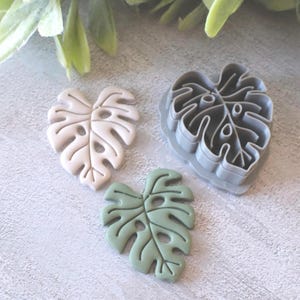 Può includere: Tre elementi a forma di foglia di monstera. Due sono foglie finite, una beige chiaro e l'altra verde tenue. Il terzo è un tagliabiscotti in plastica grigia a forma di foglia. Gli oggetti sono su una superficie grigio chiaro con fogliame verde sullo sfondo.