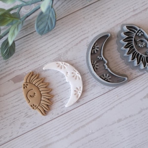 Könnte beinhalten: Vier Ausstecher im Himmelsthema: eine lächelnde Sonne, ein Halbmond mit floralen Details und zwei graue Plastikausstecher von Sonne und Mond. Die Sonne ist hellbraun und der Mond ist weiß. Die Ausstecher liegen auf einer hellen Holzmaserung.